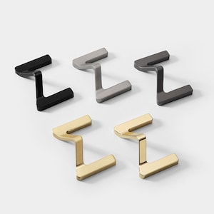 Op de afbeelding: Vijf metalen horloge standaards in verschillende afwerkingen: zwart, zilver, grijs, goud en messing. Elke standaard heeft een zigzag ontwerp.