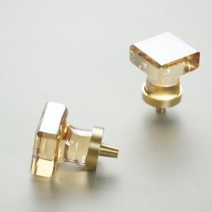 Może przedstawiać: Dwa kwadratowe szklane gałki do szafki z podstawą z pozłacanego metalu. Gałki mają przezroczysty szklany wierzch i podstawę z pozłacanego metalu.