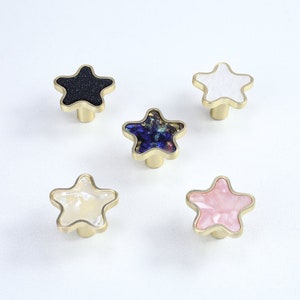 May include: Five star-shaped cabinet knobs with different coloured inlays. The knobs are made of brass and have a gold finish. The inlays are black, white, blue, cream, and pink.