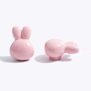 May include: Two pink ceramic bunny-shaped drawer pulls. One pull is a simple bunny shape, while the other has a more detailed design with a long ear and a small tail.