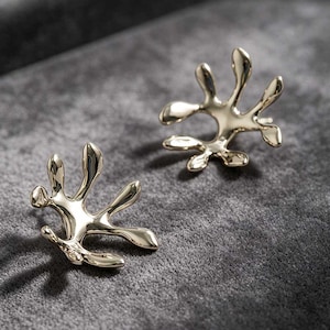 May include: A pair of gold-colored stud earrings. Each earring has an abstract, organic shape with multiple rounded, elongated points. The earrings are displayed on a gray surface, with soft lighting creating highlights and shadows.