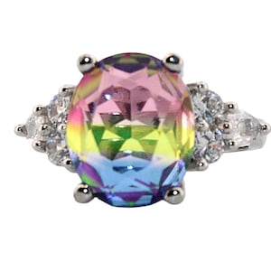 May include: A silver ring featuring a large, oval-cut gemstone with a rainbow gradient of colors. The ring is accented with smaller, clear gemstones on either side of the main stone, creating a sparkling effect. The ring is set against a white background.