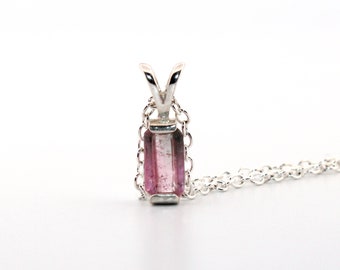 May include: A silver pendant necklace featuring a rectangular, pink and clear gemstone. The pendant is suspended from a delicate silver chain. The gemstone has a gradient of pink hues.