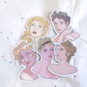 Puede incluir: Un conjunto de pegatinas troqueladas con ilustraciones de mujeres con peinados y vestimentas cl&aacute;sicas. Las pegatinas muestran diversos tonos de piel y colores de cabello, algunas adornadas con coronas de laurel. La obra de arte tiene una paleta de colores pastel suaves.