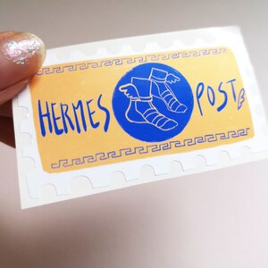 May include: A white sticker with a yellow background and a blue circle. The circle contains a drawing of a winged sandal. The text "HERMES POST" is written in blue on the yellow background.