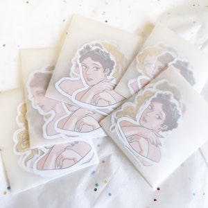 Puede incluir: Una colecci&oacute;n de pegatinas con ilustraciones de mujeres con coronas de laurel y abraz&aacute;ndose a s&iacute; mismas. Las pegatinas est&aacute;n empaquetadas en sobres blancos transl&uacute;cidos, dispuestas sobre una superficie blanca con confeti colorido.