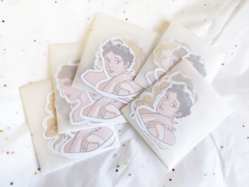 Puede incluir: Una colecci&oacute;n de pegatinas con ilustraciones de mujeres con coronas de laurel y abraz&aacute;ndose a s&iacute; mismas. Las pegatinas est&aacute;n empaquetadas en sobres blancos transl&uacute;cidos, dispuestas sobre una superficie blanca con confeti colorido.