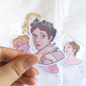 Puede incluir: Una colecci&oacute;n de pegatinas con ilustraciones de mujeres con peinados cl&aacute;sicos y coronas de flores. La pegatina central muestra a una mujer con cabello casta&ntilde;o y una expresi&oacute;n pensativa. Las pegatinas son sostenidas por una mano.