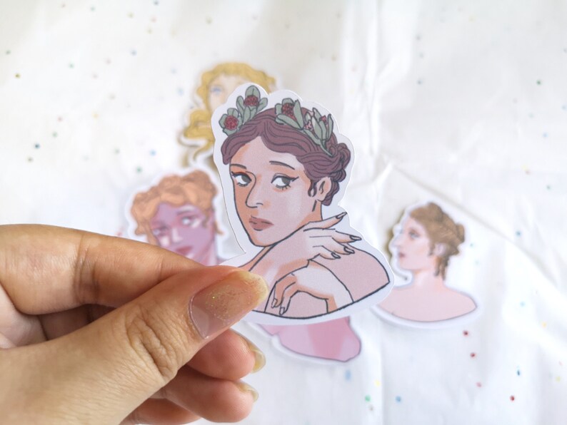 Puede incluir: Una colecci&oacute;n de pegatinas con ilustraciones de mujeres con peinados cl&aacute;sicos y coronas de flores. La pegatina central muestra a una mujer con cabello casta&ntilde;o y una expresi&oacute;n pensativa. Las pegatinas son sostenidas por una mano.