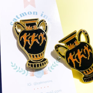 May include: Two gold-colored enamel pins shaped like ancient Greek urns. The urns are black with orange figures of people running inside. The pins are on a white background with a pink and green floral design.