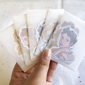 Puede incluir: Una colecci&oacute;n de pegatinas con ilustraciones de mujeres en varias poses, cada una en un paquete transparente. Las pegatinas muestran ilustraciones detalladas con paletas de colores suaves y l&iacute;neas delicadas. Las pegatinas se sostienen en una mano.