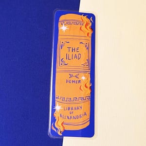 May include: A blue bookmark featuring an orange illustration of a book. The book is labelled "The Iliad" by Homer, with flames on the sides and "Library of Alexandria" at the bottom.