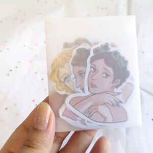 Puede incluir: Un conjunto de pegatinas con ilustraciones de mujeres con coronas de flores y retratos cl&aacute;sicos. Las pegatinas est&aacute;n en un paquete transl&uacute;cido, sostenido por una mano. Las ilustraciones tienen una paleta de colores pastel.
