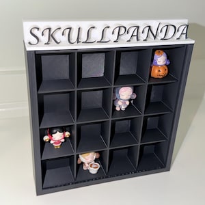 May include: A black square display shelf with sixteen individual compartments, topped with a white horizontal bar featuring the black text SKULLPANDA. Four small stylized figurines are placed within the grid, including one sitting on a pumpkin.