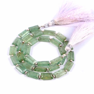 May include: A necklace of light green, rectangular beads with silver spacers. The beads are strung together. White tassels with silver detailing are attached to each end of the necklace.