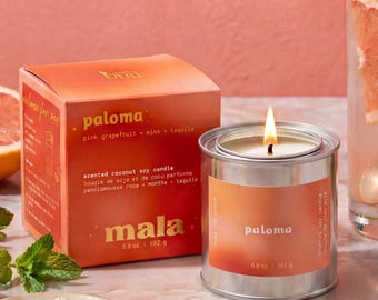 Könnte beinhalten: Eine Duftkerze aus Kokos-Soja-Wachs in einer silbernen Dose mit dem Etikett 'paloma' und '6.8 oz | 192 g', begleitet von der orangefarbenen Schachtel, einem Grapefruit-Cocktail mit Salzrand, Grapefruitscheiben, Minzblättern und Meersalz.
