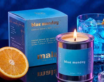 Könnte beinhalten: Eine Metalldose mit einer Kerze, beschriftet mit Blue Monday und einer hellblauen Papierbanderole, mit brennendem Docht. Daneben stehen eine passende hellblaue Pappschachtel, eine halbierte Orange und ein Glas mit blauem Getränk und Eiswürfeln.
