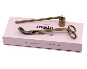 Könnte beinhalten: Ein Kerzenpflege-Set in Messingoptik mit einem Dochtschneider und einem glockenförmigen Kerzenlöscher auf einer hellrosa rechteckigen Schachtel. Die Schachtel ist mit dem schwarzen Text "mala" und "because your candle deserves self-care too, babe" bedruckt.