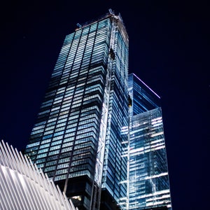 May include: A view of two tall, modern skyscrapers with glass facades illuminated at night. The buildings are located in a city with a white, geometric structure in the foreground.