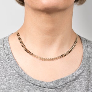 Op de afbeelding: Een gouden ketting met een chevron-patroon. De ketting is gemaakt van kleine, met elkaar verbonden stukjes die een doorlopende pijlvorm vormen.