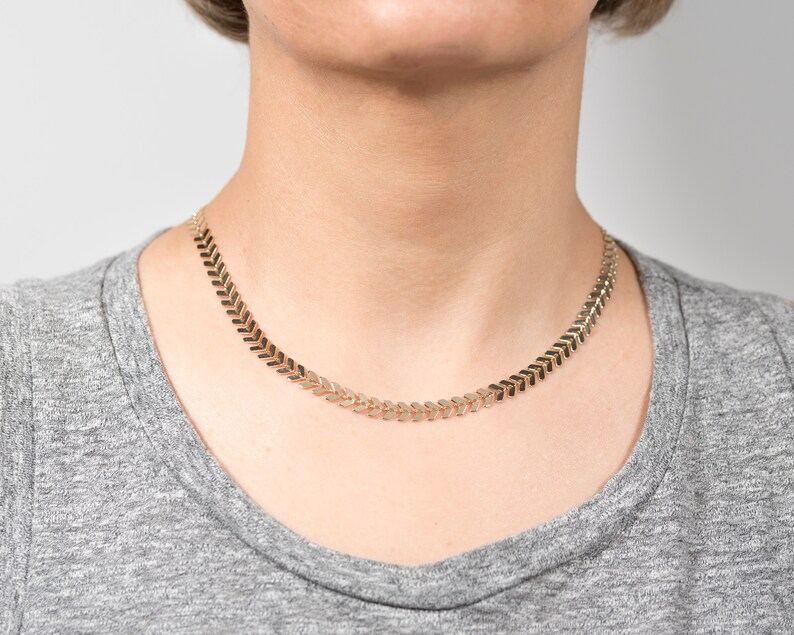 Op de afbeelding: Een gouden ketting met een chevron-patroon. De ketting is gemaakt van kleine, met elkaar verbonden stukjes die een doorlopende pijlvorm vormen.