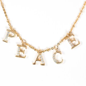 Puede incluir: Un collar de oro con la palabra "PEACE" escrita en letras individuales. Cada letra está adornada con pequeñas piedras de circonita cúbica transparentes.