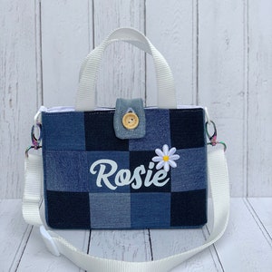 May include: A small blue denim tote bag with a white daisy and the name "Rosie" embroidered on the front. The bag has a white strap and a top handle.