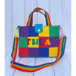 May include: A small rainbow-coloured tote bag with a patchwork design and the name "THEA" in large, colourful letters. The bag has a top handle and a long adjustable strap.