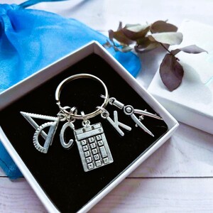 May include: A silver-toned metal keyring featuring four charms: a protractor, the letter K, a calculator, and a drafting compass. The keyring is presented inside a white square gift box with a black velvet insert, accompanied by a blue fabric pouch.