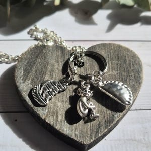 May include: A silver necklace featuring a feather, a seated imp, and a half-circle charm. The necklace is displayed on a heart-shaped wooden board. The charms are attached to a silver chain.