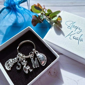 May include: A silver keyring with charms including a flip-flop, a cat, and the letters "S" and "M". The keyring is in a white box with the words "Sleepy Koala" on the lid. A blue drawstring bag is visible in the background.