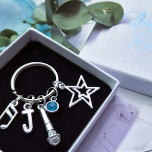 May include: A silver keyring with a star, musical notes, a microphone, and a letter J charm. The keyring is in a white gift box with the words "Always A Music" printed on the top.