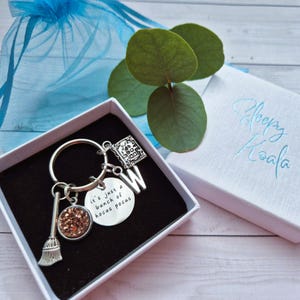 May include: A silver keychain featuring a broom charm, a round charm with the text "it's just a bunch of hocus pocus", a book charm, and the letter "W". The keychain is presented in a white box with the text "Sleepy Koala".