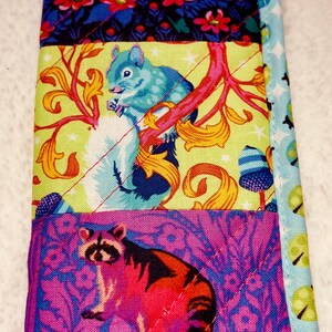 May include: A quilted fabric item featuring three distinct patterned sections. The top section has a blue background with pink flowers. The middle section showcases a blue squirrel on a yellow background. The bottom section displays a raccoon on a purple background.