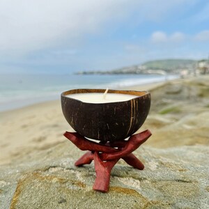 May include: A white coconut shell candle with a wooden wick sits on a red wooden stand. The candle is on a sandy beach with the ocean in the background.