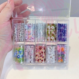 May include: A clear plastic storage box with 10 rolls of nail art transfer foil. The foil has various floral patterns in shades of pink, purple, blue, green, and white.