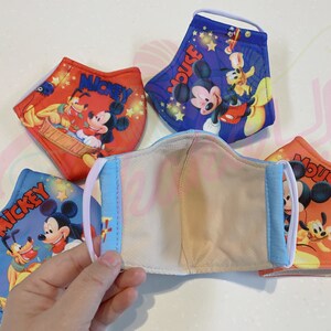 K&ouml;nnte beinhalten: Vier wiederverwendbare Gesichtsmasken mit Mickey Mouse und Pluto. Die Masken bestehen aus einem weichen, atmungsaktiven Stoff und haben verstellbare Ohrschlaufen. Die Masken sind mit verschiedenen bunten Designs bedruckt, darunter Sterne, Mickeys Unterschrift und die Worte "Mickey Mouse".