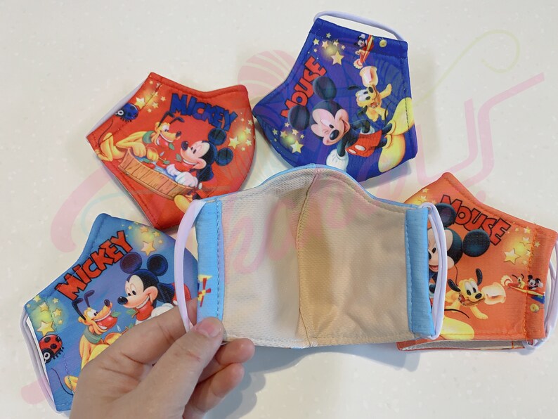 K&ouml;nnte beinhalten: Vier wiederverwendbare Gesichtsmasken mit Mickey Mouse und Pluto. Die Masken bestehen aus einem weichen, atmungsaktiven Stoff und haben verstellbare Ohrschlaufen. Die Masken sind mit verschiedenen bunten Designs bedruckt, darunter Sterne, Mickeys Unterschrift und die Worte "Mickey Mouse".