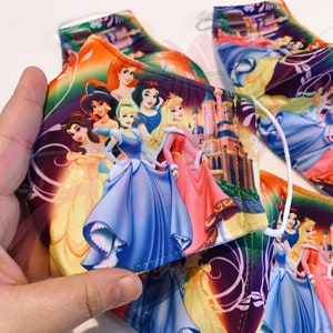May include: A set of colourful face masks featuring Disney princesses, including Cinderella, Snow White, Belle, and Aurora. The masks are made of fabric and have a purple background with floral designs.