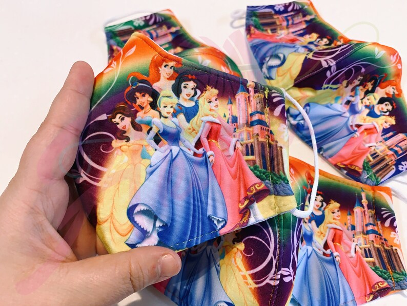May include: A set of colourful face masks featuring Disney princesses, including Cinderella, Snow White, Belle, and Aurora. The masks are made of fabric and have a purple background with floral designs.
