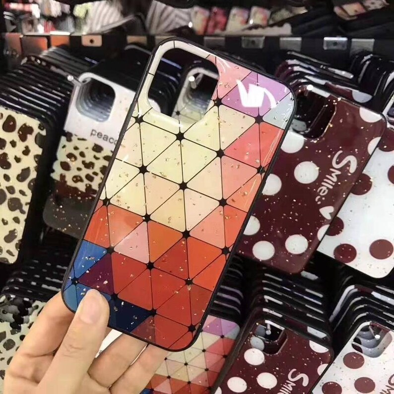 May include: A collection of colorful phone cases with various designs, including geometric patterns, polka dots, and animal prints. Some cases feature gold glitter accents. The cases are displayed on a shelf.