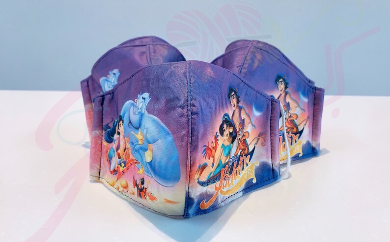 May include: Three purple face masks featuring a printed image of Aladdin, Jasmine, Abu, and Genie from the Disney film Aladdin. The masks have white elastic ear loops.