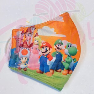 May include: A reusable face mask with a colorful print of Mario, Luigi, Peach, Toad, and Yoshi characters from the Super Mario Bros. video game series. The mask has a peach background with a castle and clouds.