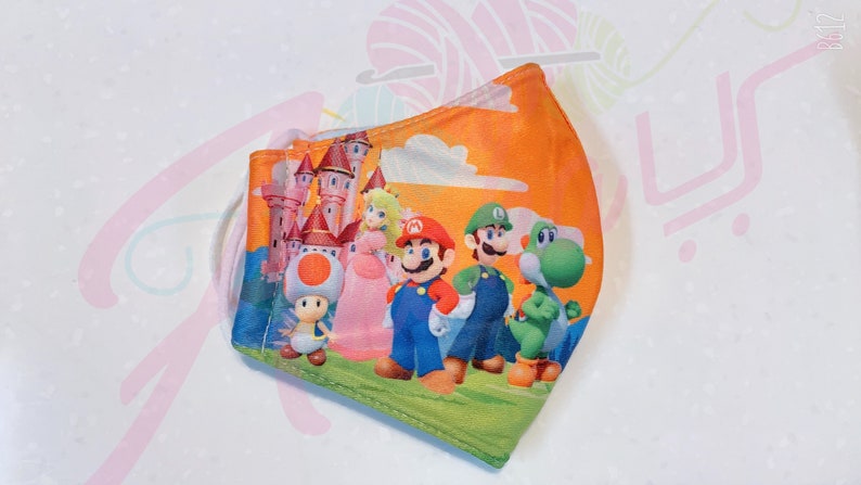 May include: A reusable face mask with a colorful print of Mario, Luigi, Peach, Toad, and Yoshi characters from the Super Mario Bros. video game series. The mask has a peach background with a castle and clouds.