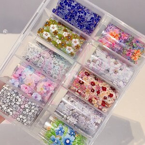 May include: A clear plastic box filled with ten rolls of nail art transfer foil. Each roll features a different floral design with various colors, including pink, blue, green, white, and black.