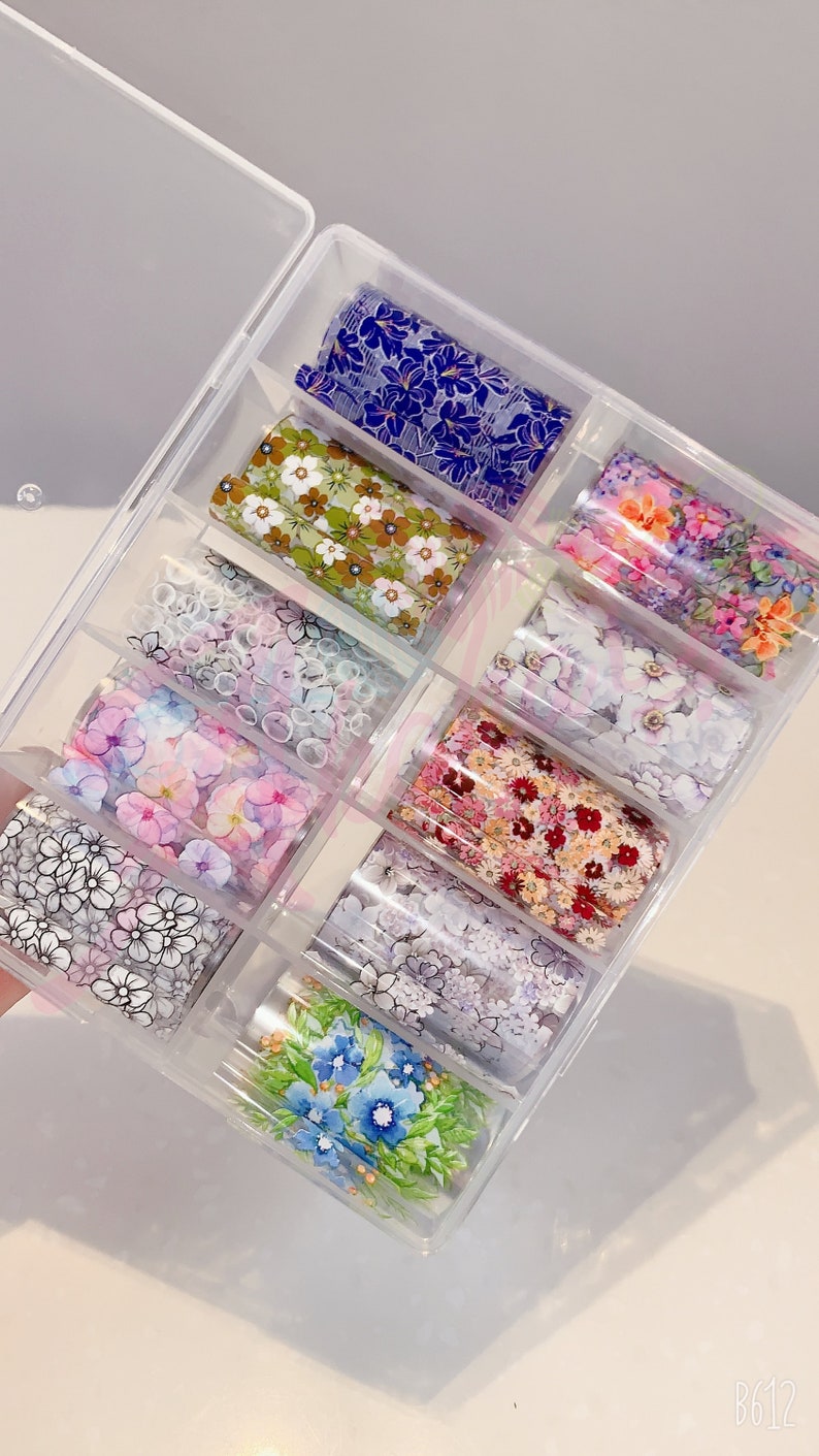 May include: A clear plastic box filled with ten rolls of nail art transfer foil. Each roll features a different floral design with various colors, including pink, blue, green, white, and black.