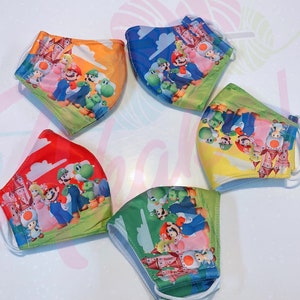 May include: Five colorful face masks with a cartoon image of Mario and friends from the Super Mario Bros. video game series. The masks are made of fabric and have elastic ear loops.