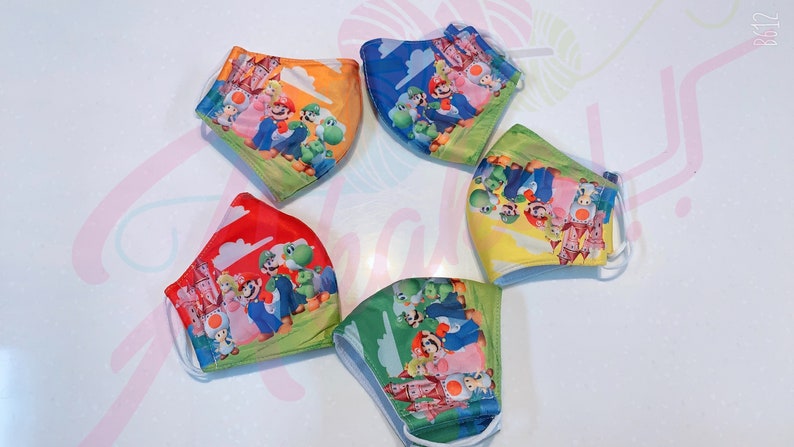 May include: Five colorful face masks with a cartoon image of Mario and friends from the Super Mario Bros. video game series. The masks are made of fabric and have elastic ear loops.