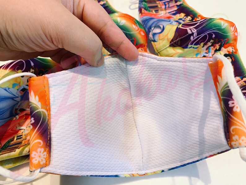 May include: A white face mask with a colourful Disney Princesses print. The mask has a white inner layer and orange elastic straps.
