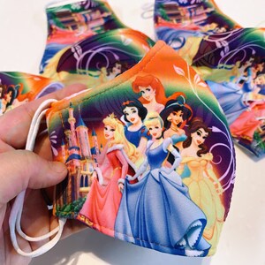 May include: A colourful face mask featuring a group of Disney princesses, including Cinderella, Snow White, Belle, and Ariel. The mask has a purple, orange, and green background with a castle in the background.